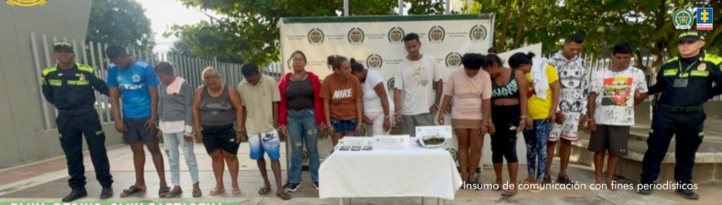 En la fotografía aparecen varias personas entre hombres y mujeres de pie, de frente con los brazos esposados, custodiados por dos uniformados de la policía Nacional. Detrás de ellos hay un pendón de esa institución.