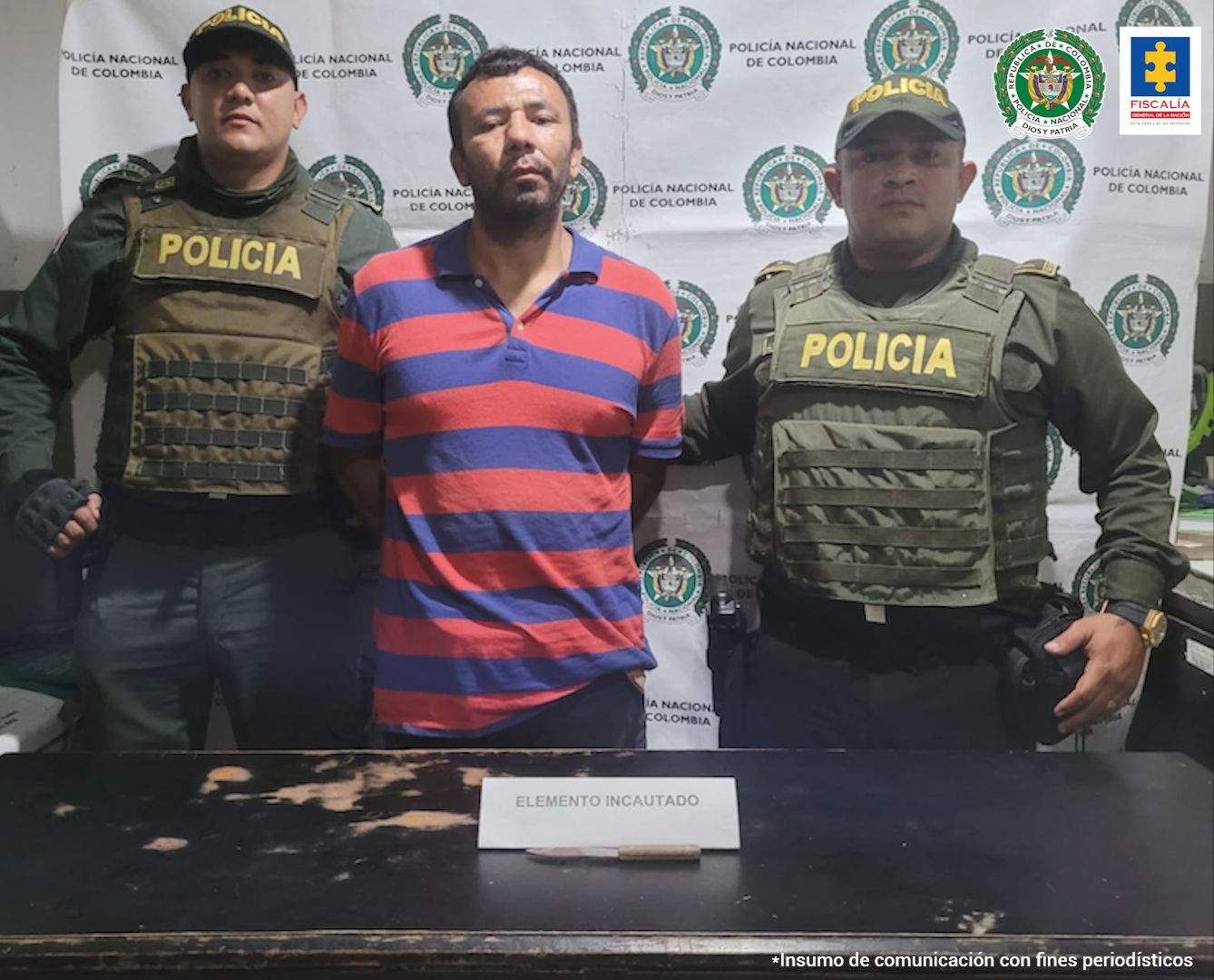 En la fotografía se observa a un hombre con camiseta a rallas rojas con azul custodiado por dos agentes de la Policía Nacional, delante de un pendón de esa institución.