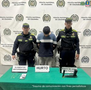 En la fotografía está un hombre trigueño, de pie, de frente, con la cabeza agachada, cabellos negros, los brazos atrás esposados. Está vestido con un buzo de rayas azul, blanco y negro y un jean azul claro. Se encuentra custodiado por dos uniformados de la Policía Nacional y delante de ellos hay una mesa con un arma de fuego y un celular.