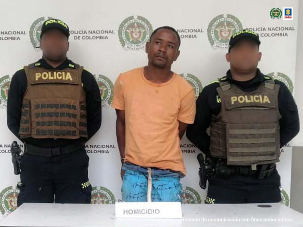 En la fotografía aparece un hombre moreno, de pie, con los brazos atrás, vestido de camiseta naranja y jean azul, custodiado por dos agentes de la Policía Nacional. Detrás de ellos hay un pendón de esa institución.