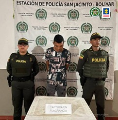 En la imagen se observa a un hombre con camiseta negra y pantalón negro con la cabeza agachada, custodiado por dos agentes de la Policía Nacional delante de un pendón de esa institución.