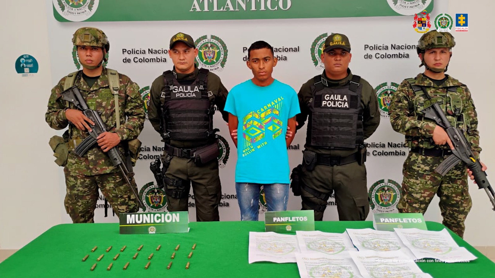 En la fotografía está un hombre de pie, joven, con los brazos atrás esposados, custodiado por uniformados de la Policía y Ejército Nacional. Se encuentra vestido con una camiseta y jean azul.
