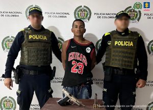En la imagen aparece un hombre moreno, delgado, de mediana estatura, vestido con una camisilla negra con roja y un jean azul. Se encuentra custodiado por dos policías y delante de ellos hay una mesa con el arma de fuego que se le incautó.