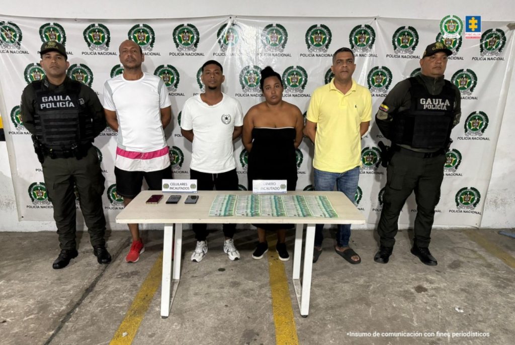En la fotografía están tres hombres y una mujer de pie, de frente, con los brazos esposados custodiado por dos uniformados de la Policía Nacional. Se encuentran acompañados por uniformados de la Policía Nacional y delante de ellos hay una mesa donde está el dinero incautado.