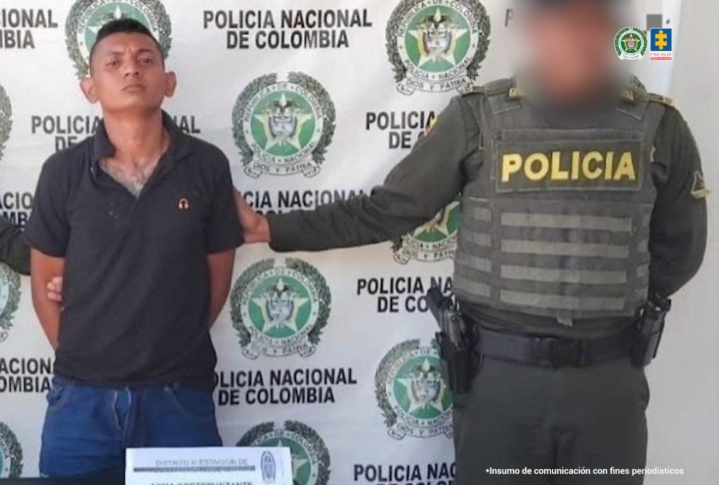 En la foto se encuentra un hombre moreno de pie, con los brazos atrás esposados, de cabellos cortos negros, vestido con camiseta negra y jean azul. Se encuentra custodiado por un uniformado de la Policía Nacional. 