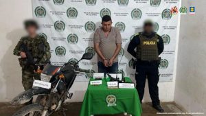 n la imagen está un hombre de pie, con los brazos hacia adelante esposado, con la cabeza agachada, pelo negro, trigueño, vestido con una camiseta marrón y un jean azul, custodiado por un uniformado de la Policía Nacional y por otro del Ejército. Delante de ellos hay una mesa con las armas incautadas.