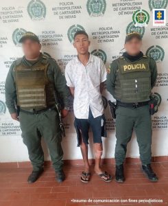En la imagen aparece un hombre con brazos atrás, vestido de camisa blanca con bermuda azul oscura y sandalias negras, custodiados por dos uniformados de la Policía Nacional. Detrás de ellos hay un pendón de esa institución.