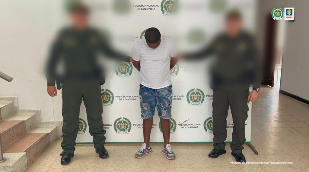 En la imagen se observa a un hombre de camiseta blanca con bermuda de jean y tenis, custodiado por dos agentes de la Policía Nacional delante de un pendón de esa institución.