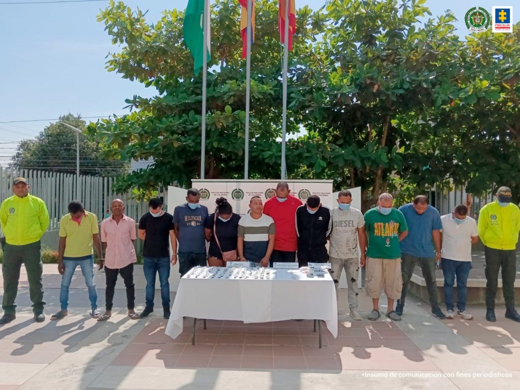 En la fotografía se observan a doce personas custodiadas por dos agentes de la Policía Nacional. Delante de ellos se encuentra una mesa blanca que muestra la droga que les fue incautada durante sus capturas.