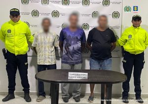 En la fotografía, están tres hombres de pie, con los brazos atrás, sus rostros difusos, custodiados por dos uniformados de la Policía Nacional, delante de un pendón de esa institución.
