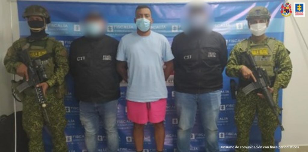 En la imagen se observa a un hombre de camiseta gris con bermuda rosada custodiado por dos uniformados del CTI y dos del Ejército Nacional, delante de un pendón de la Fiscalía General de la Nación.