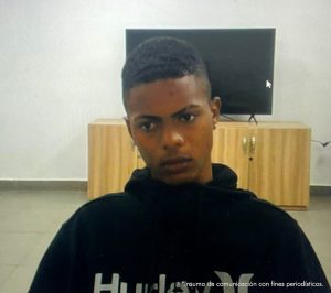 En la imagen se ve el medio cuerpo de un hombre joven moreno, ojos y nariz mediana, labios gruesos, cabellos cortos negros, vestido con un buzo negro. Se encuentra sentado en una silla asistiendo virtualmente a la audiencia en la que fue procesado.