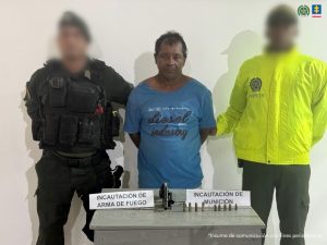 En la fotografía aparece un hombre de mediana edad y estatura, moreno, de pie, con los brazos atrás, vestido con una camiseta azul, custodiado por dos uniformados de la Policía Nacional. Delante de ellos hay una mesa con el arma y las municiones incautadas.