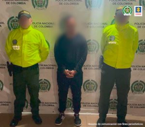 En la fotografía se encuentra un hombre de pie, con las manos esposadas, rostro difuso, vestido de buzo, sudadera y tenis negros custodiado por dos uniformados de la Policía Nacional.