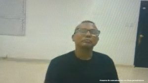 En la fotografía aparece un hombre moreno con gafas, de edad madura, cabellos cortos, vestido con camiseta negra. Se encuentra sentado en el interior de la sala de audiencia donde se le procesó. 