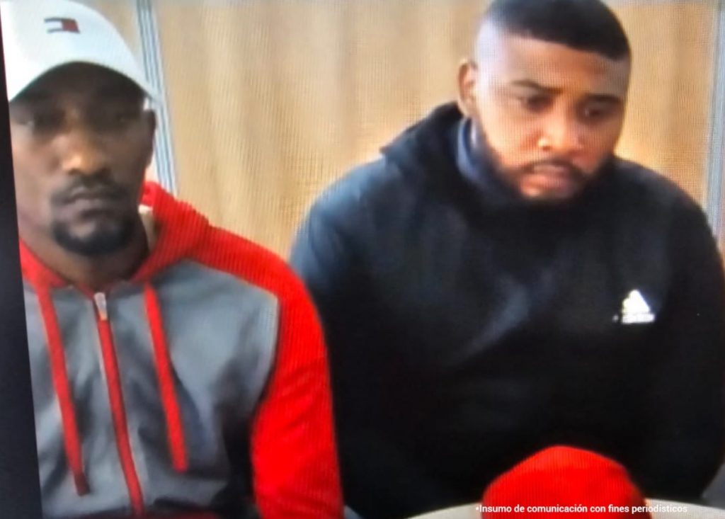 En la imagen se ve los medios cuerpos de dos hombres morenos, de contextura mediana, ambos de barbas, uno vestido de buzo rojo con gris y gorra blanca y el otro usa un buzo azul oscuro. Se encuentran sentados asistiendo virtualmente a la audiencia que los procesó.