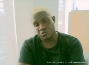 En la imagen está el medio cuerpo de un hombre moreno, de contextura gruesa, facciones faciales gruesas, cabellos crespos con tinte amarillo, se encuentra sentado con los brazos cruzados asistiendo virtualmente a la audiencia en la que se le procesó.