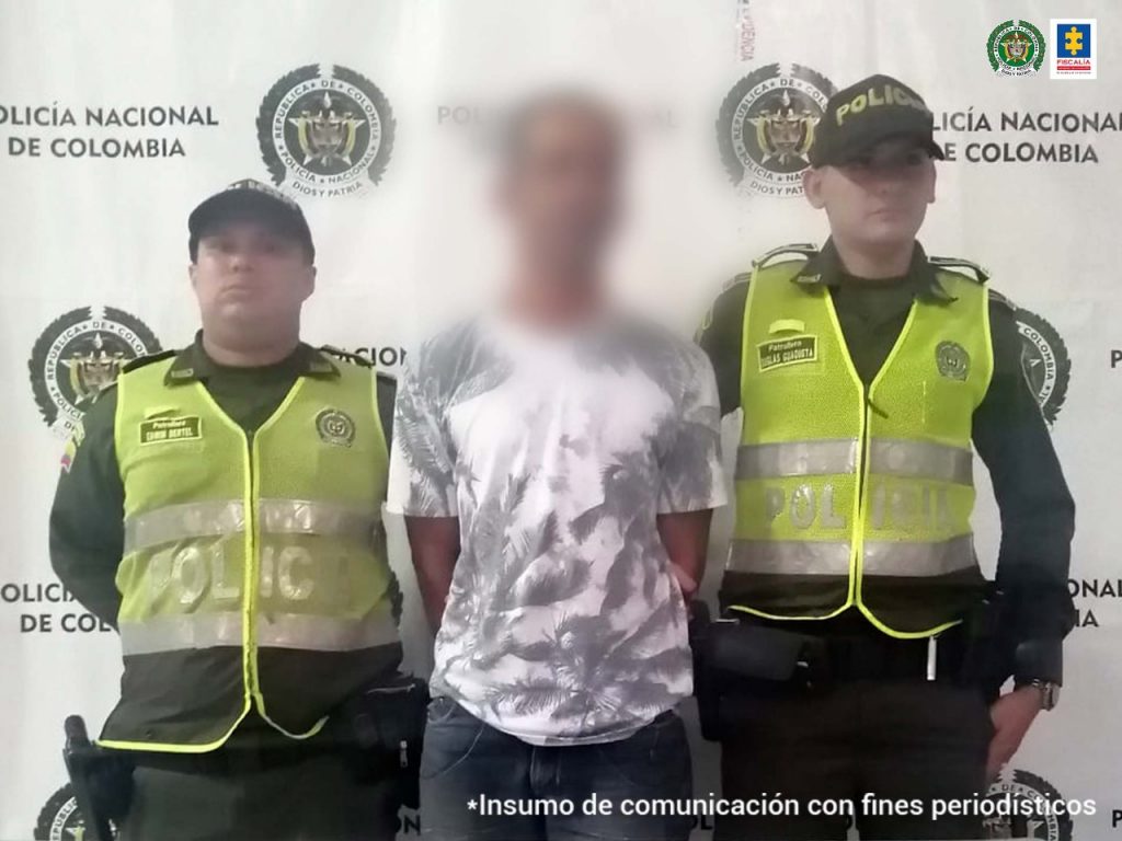 En la fotografía se observa a un hombre con una camiseta blanca, rostro difuminado, custodiado por dos policías delante de un pendón de esa institución.