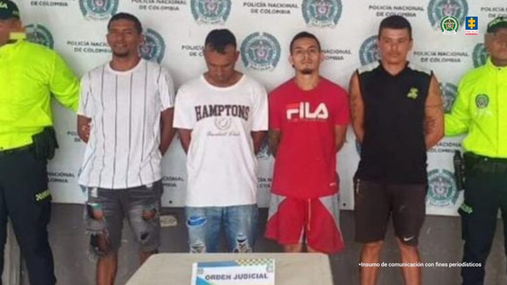 En la fotografía aparecen cuatro hombres de pie, con los brazos atrás esposados, morenos, custodiados por dos uniformados de la Policía Nacional. Detrás de ellos hay un pendón de esa institución.