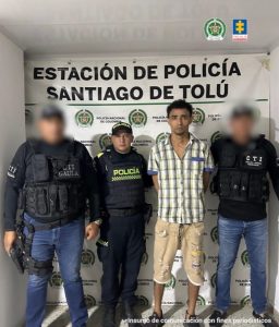 En la fotografía aparece un hombre alto de pie, delgado, con los brazos atrás, trigueño, cabellos negros, vestido con camisa manga corta de cuadros amarillos, gris y azul, bermuda beige y unos zapatos negros custodiado por uniformados del CTI y la Policía Nacional.