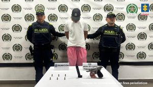 En la fotografía, está un hombre de pie, de frente, con los brazos atrás esposados, contextura delgada, moreno, con la cabeza agachada, tiene una gorra negra y está vestido con camiseta blanca y bermuda rosada. Se encuentra custodiado por dos uniformados de la Policía Nacional.