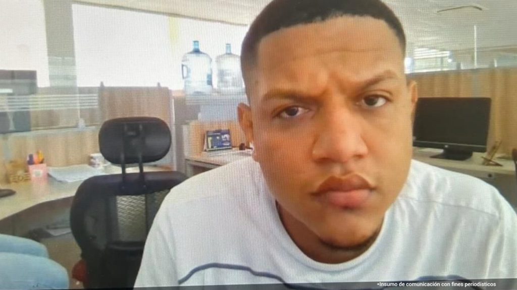 En la fotografía está el rostro de un hombre trigueño, cara redonda, con labios y nariz gruesos, cabellos cortos crespos negros, vestido con una camiseta blanca. Se encuentra sentando en una oficina asistiendo de manera virtual a la audiencia en la que se le procesó.
