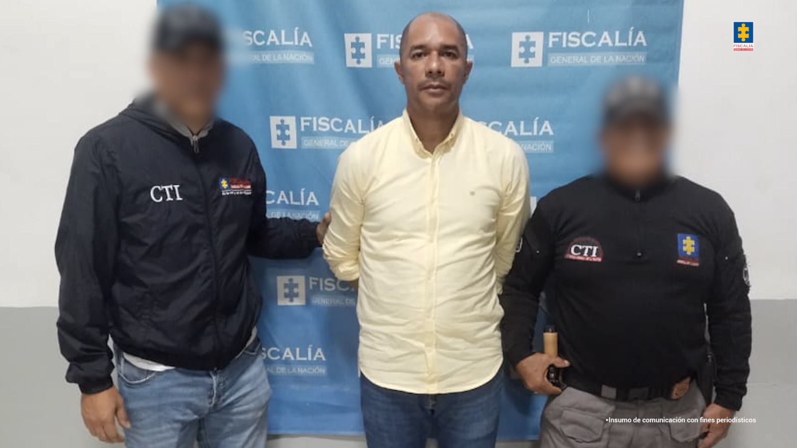 En la fotografía está un hombre moreno de pie, semi calvo, con los brazos atrás, vestido con una camisa manga larga amarilla y un jean azul, custodiado por dos uniformados del CTI. Detrás de ellos hay un pendón de la Fiscalía.