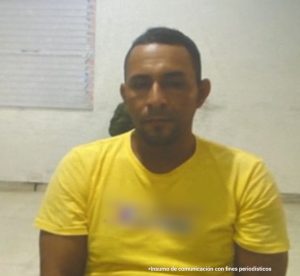 En la imagen está el medio cuerpo de un hombre, moreno, de cabellos cortos, semi barbado, está sentado en una silla, vestido con camiseta amarilla manga corta asistiendo de manera virtual a la audiencia en la que se le procesó.
