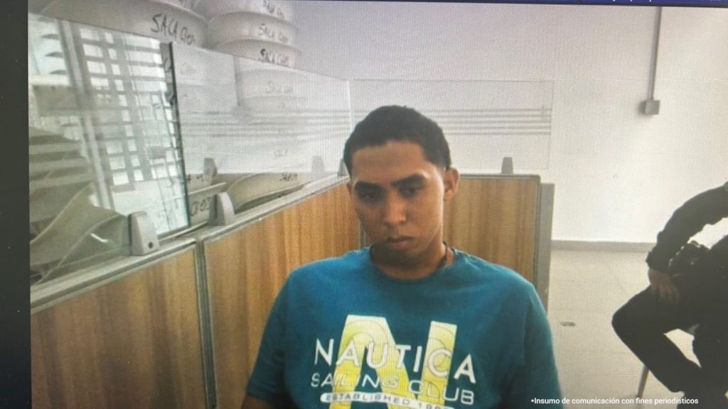 En la imagen está el medio cuerpo de un hombre moreno, con cejas abundantes, cabellos negros, ojos, nariz y boca mediana, vestido con una camiseta azul que tiene letras amarillas. Se encuentra sentado en una silla dentro de una oficina asistiendo virtualmente a la audiencia en la que se le procesó.