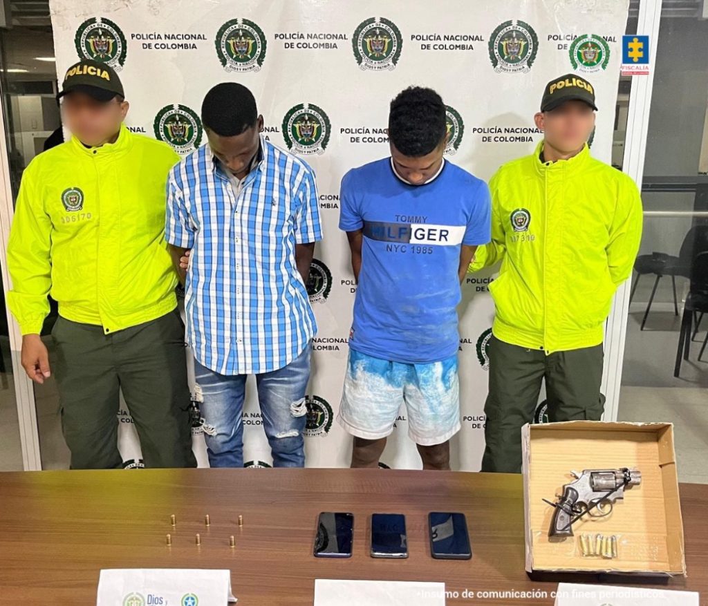En la fotografía aparecen dos hombres morenos, delgados, de pie, con los brazos atrás esposados. Uno se encuentra vestido con camisa de cuadros azul con blanco y jean azul y el otro con camiseta azul y bermuda azul con blanca. Están custodiados por dos agentes de la Policía Nacional.