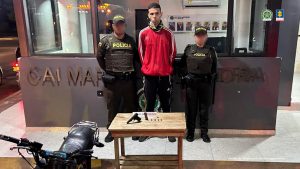  En la foto aparece un hombre de pie, alto, con los brazos atrás, vestido de buzo rojo y pantalón negro, custodiado por dos agentes de la Policía Nacional. Delante de ellos está una mesa con el arma que le incautó.