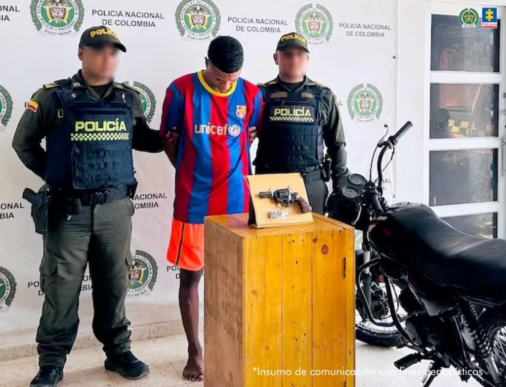En la fotografía está un hombre moreno de pie, con los brazos atrás esposados, cabeza agachada de cabellos negros crespos, está vestido con una camiseta de rayas rojas con azul oscuro y una bermuda naranja. Se encuentra custodiado por dos uniformados de la Policía Nacional.