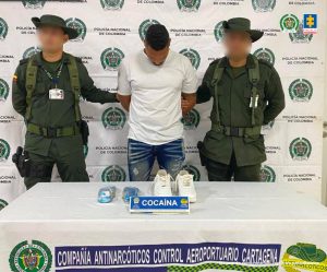 En la fotografía hay un hombre de pie con los brazos atrás, vestido de camiseta blanca con jean azul, custodiado por dos agentes de la Policía Nacional. Delante de ellos está la droga incautada y detrás de ellos hay un pendón de la Policía.