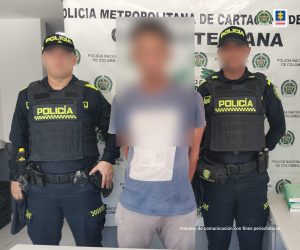En la fotografía aparece un hombre de pie, con los brazos atrás, el rostro difuso, vestido de camiseta gris con pantalón beige, custodiado por dos agentes de la Policía Nacional. Detrás de ellos hay un pendón de esa institución.