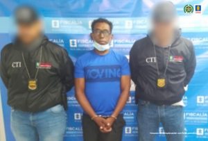 En la imagen se observa a un hombre moreno con gafas, vestido con una camiseta azul custodiado por dos uniformados del CTI de la Fiscalía, delante de un pendón de esa institución.