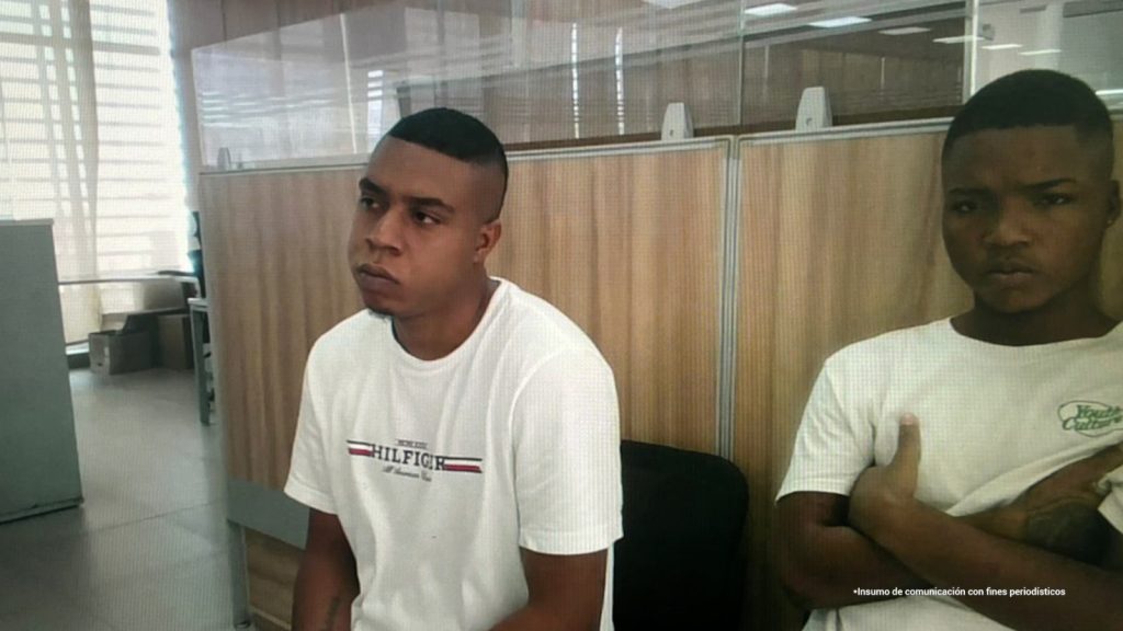 En la fotografía están dos hombres jóvenes, morenos de cabellos cortos negros, ambos vestidos con camiseta blanca, sentados al interior de una oficina asistiendo virtualmente a la audiencia en la que se les procesó.