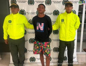  En la foto aparece un hombre de pie, moreno, de cabellos crespos negros corto, con los brazos atrás esposado, vestido con una camiseta negra y bermuda estampada de colores, custodiado por dos agentes de la Policía Nacional.
