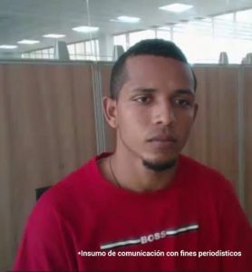 En la foto está un hombre joven, moreno, cabellos negros crespos cortos, tiene una barba pequeña debajo del mentón, está vestido con una camiseta roja y se encuentra al interior de una oficina asistiendo virtualmente a la audiencia en la que se le procesó.