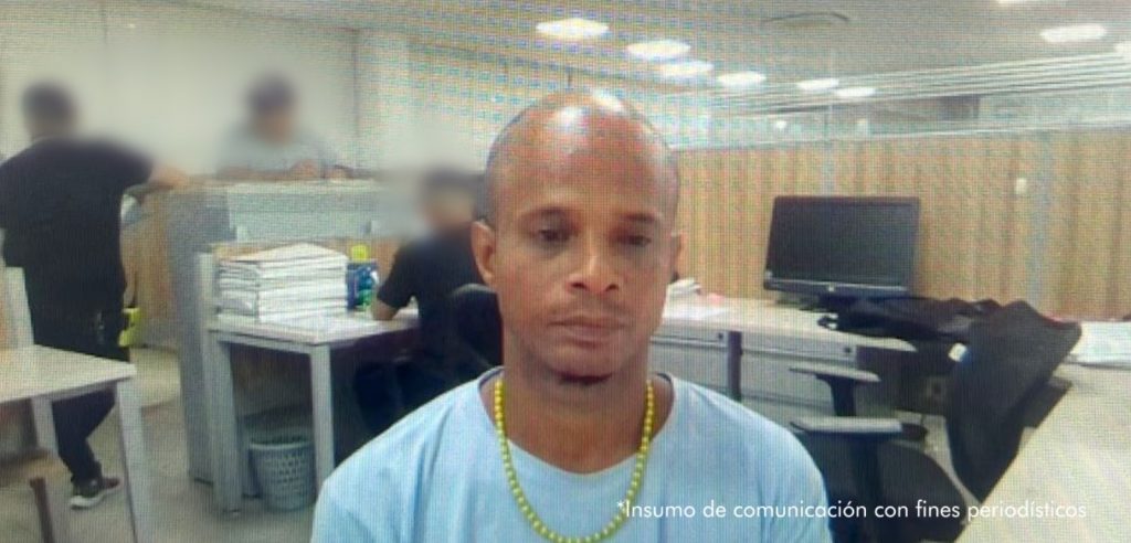 En la fotografía está el medio cuerpo de un hombre trigueño, calvo, delgado, vestido con una camiseta azul y un collar de pepas verde. Se encontraba sentado al interior de una oficina donde asistía virtualmente a la audiencia en la que se le procesó.