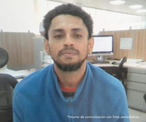 En la fotografía aparece el medio cuerpo de un hombre joven, trigueño, cabellos negros, con semi barba, aretes, con ojos, nariz, orejas y boca grande, vestido con una camiseta azul. Se encuentra sentado en una oficina escuchando virtualmente la audiencia en la que se le procesó. 