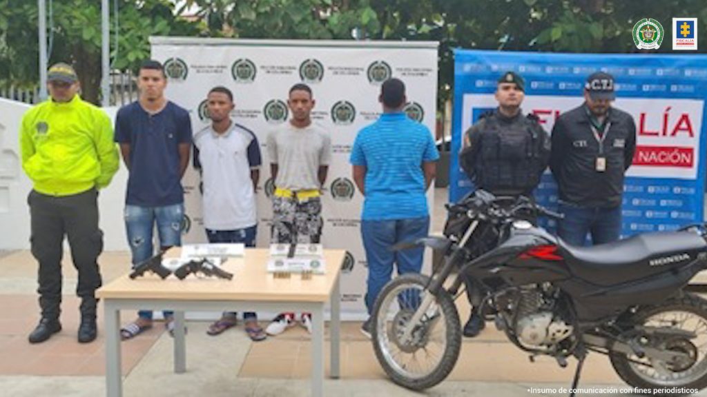 En la fotografía se observan a tres hombres de frente y a uno de espalda custodiado por uniformados del CTI y Policía Nacional. Delante de ellos aparece una mesa con arma de fuego, municiones y la motocicleta que les fueron incautados.
