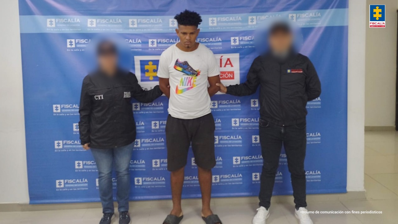 En la foto está un hombre joven, delgado, de pie, de frente, alto, moreno, tiene el cabello negro crespo, largo, tipo afro, está vestido con camiseta blanca, bermuda y sandalias negras. Se encuentra custodiado por dos uniformados del Cuerpo Técnico de Investigación (CTI).