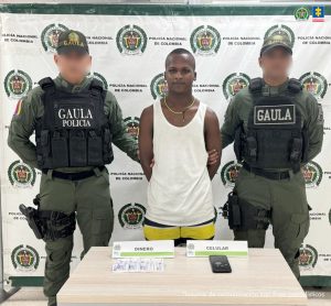 En la fotografía aparece un hombre moreno de pie, con los brazos atrás, Vestido de camisilla blanca y bermuda amarilla, con blanco y negro, custodiado por dos uniformados de la Policía Nacional. Delante de ellos hay una mesa con dinero y un celular incautado.