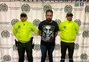 En la imagen se ve a un hombre, identificado como Sebastián Cano Ospina, esposado y vestido con ropa casual, escoltado por dos policías uniformados que lo sujetan de los brazos.
