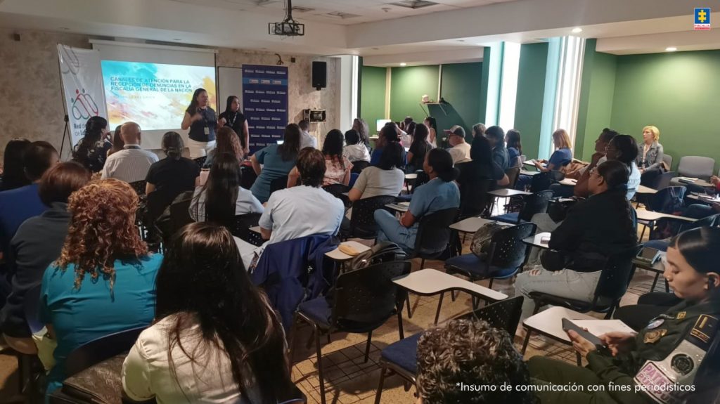 Las fotografías fueron tomadas dentro de un auditorio donde hay un grupo grande de personas, sentadas en pupitres de capacitación, apreciando una proyección en una pantalla que se encuentra en el fondo. A los extremos hay dos pendones institucionales de la Fiscalía y de la Red de Salud de Ladera (ESE). Asimismo, hay una servidora de la Fiscalía explicando lo que se proyecta en la pantalla.