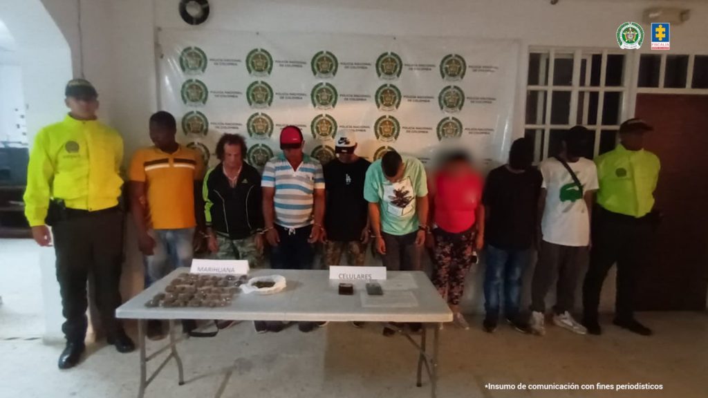 En la fotografía se aprecia a siete presuntos integrantes de una banda delincuencial dedicada al narcomenudeo en Caicedonia (Valle del Cauca). A los extremos de ellos hay uniformados de la Policía Nacional. Delante de ellos hay una mesa con mantel verde y sobre ella hay bolsas plásticas transparentes y pequeñas con contenido verdoso, al igual que dos teléfonos celulares. Asimismo, hay dos letreros que dicen: ‘Marihuana’, y ‘Celulares’. Detrás de ellos hay un pendón institucional de la Policía.