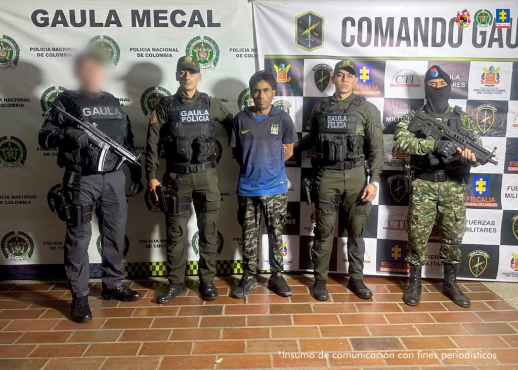 En la fotografía se aprecia de pie y con los brazos atrás a Brayan Eduardo Garcés Peláez. Viste camiseta azul con estampado negro y gris, pantalón camuflado y zapatillas negras. A su extremo derecho hay un servidor del CTI de la Fiscalía y del Gaula Policía y al extremo izquierdo un uniformado del Gaula Policía y del Ejército. Detrás de ellos hay dos pendones institucionales de la Policía Nacional y otro del Comando Gaula con logos de la Fiscalía y del Ejército.