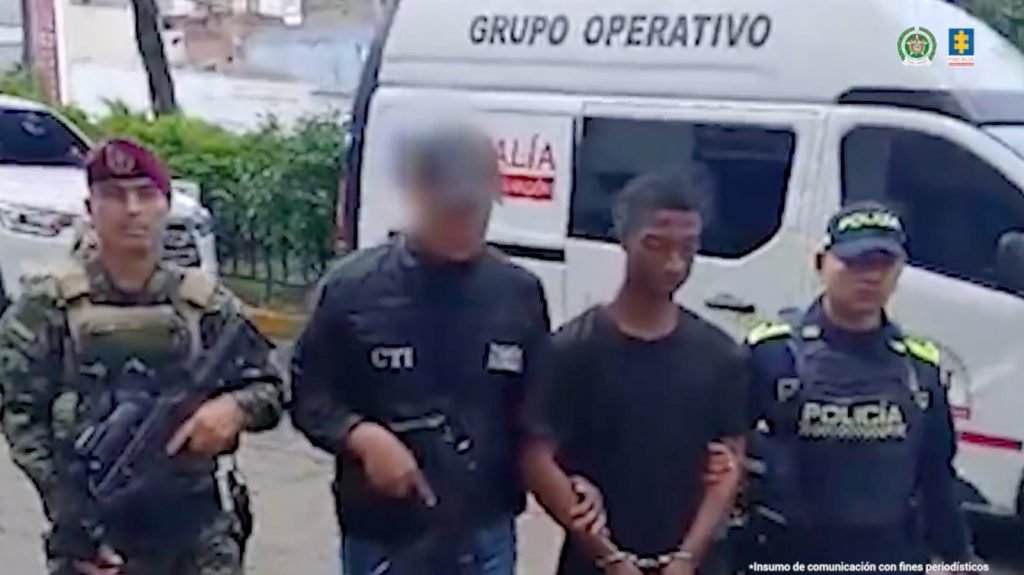 La fotografía está tomada en exteriores y se aprecia en acción de caminata, con los brazos adelante y esposado a Deivy Alexander Arboleda Delgado. Viste camiseta negra de manga corta. A su derecha hay un servidor del CTI de la Fiscalía y un uniformado del Ejército; a la izquierda un uniformado de la Policía Nacional. Atrás de ellos hay un vehículo institucional de la Fiscalía.