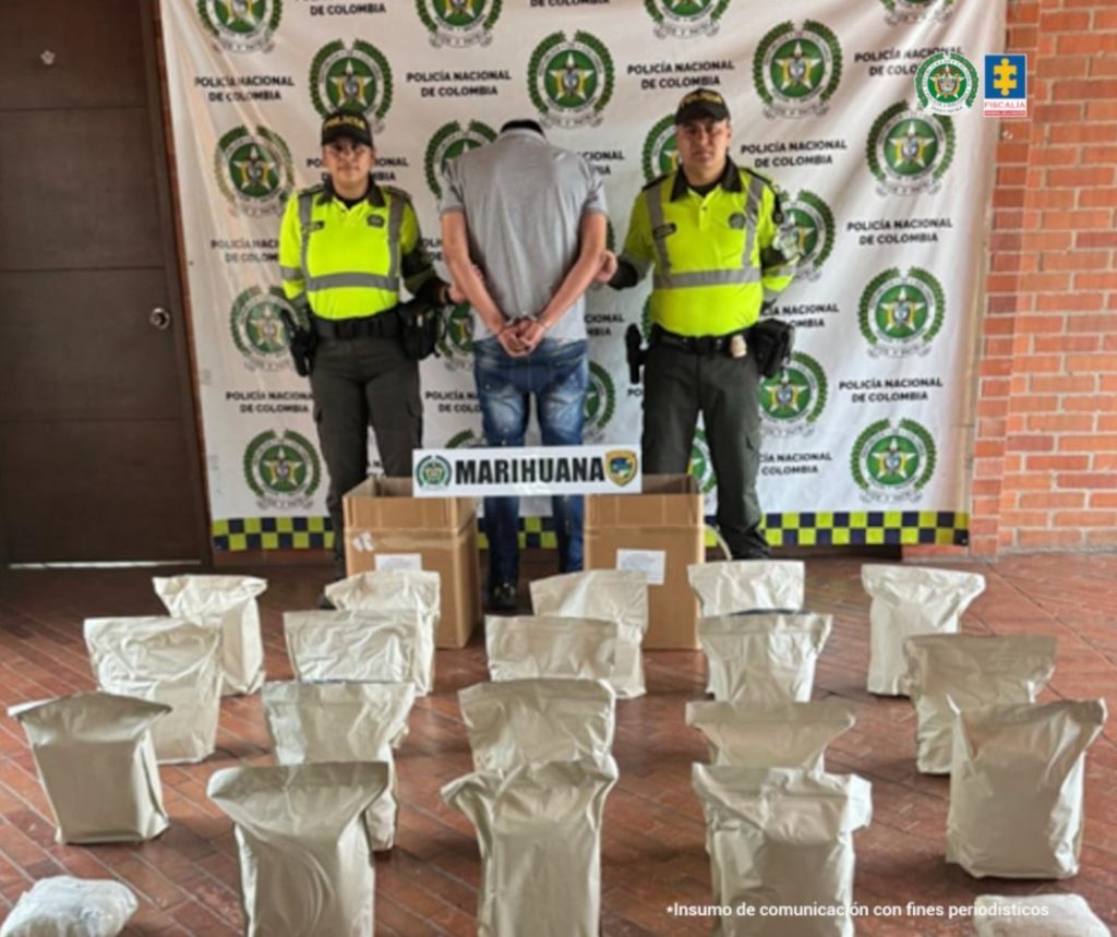 En la fotografía se aprecia de pie, de espaldas a la cámara, los brazos atrás y esposado a Deybi Hernán Gaviria Gaviria. Viste con camisa de color gris y mangas cortas; jean azul desgastado. A sus extremos hay uniformados de la Policía Nacional. En el suelo hay dos cajas de cartón y sobre ellas un letrero, con logos de la Policía, que dice: ‘Marihuana’. Asimismo hay 20 bolsas blancas. Detrás del judicializado y los uniformados hay un pendón institucional de la Policía Nacional.