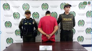 En la fotografía se aprecia de espaldas a la cámara, con los brazos atrás y esposado a Diego Edinson Trujillo Tobón. Viste camiseta de manga corta de color rojo. A lado y lado de él hay uniformadas de la Policía Nacional. Delante de ellos hay una superficie marrón y sobre ella un letrero que dice: ‘Violencia intrafamiliar’. Detrás de ellos hay un pendón institucional de la Policía.